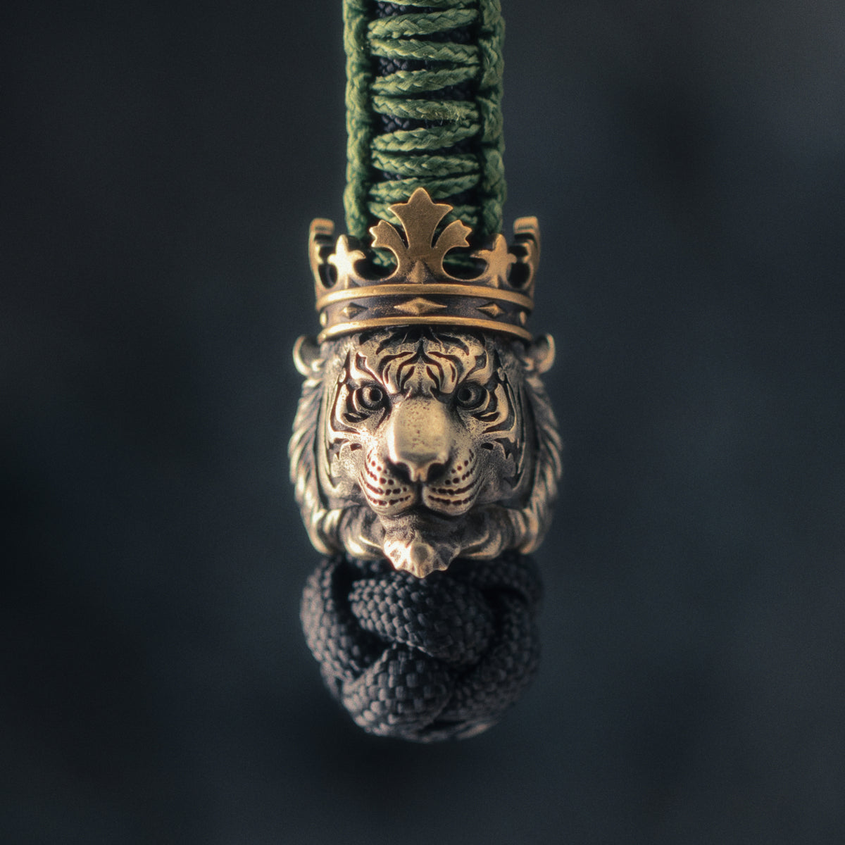 Front view of a Tiger King EDC bead attached to green and black paracord, showing the sculpted tiger face and antique brass crown.