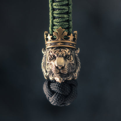 Front view of a Tiger King EDC bead attached to green and black paracord, showing the sculpted tiger face and antique brass crown.