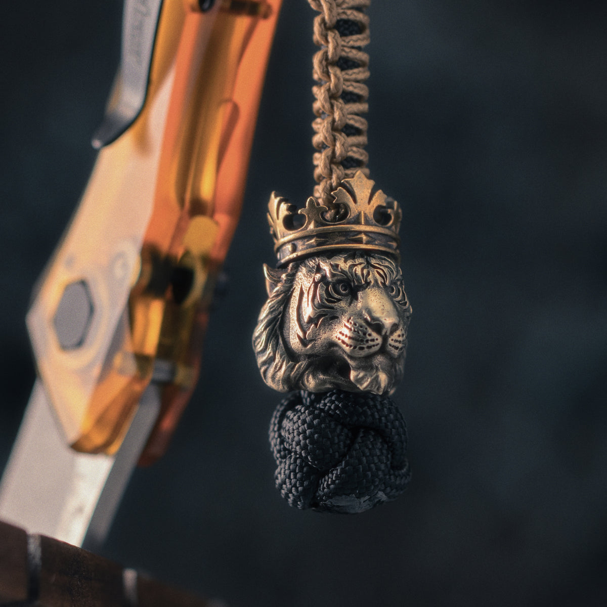 Crowned tiger EDC bead hanging beside a folding knife, showing engraved tiger stripes, sculpted fur and hand-aged metal surfaces.