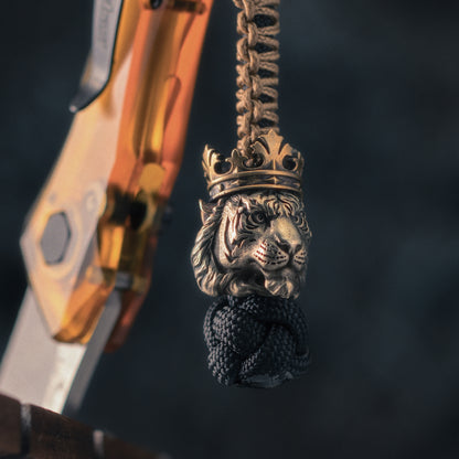 Crowned tiger EDC bead hanging beside a folding knife, showing engraved tiger stripes, sculpted fur and hand-aged metal surfaces.