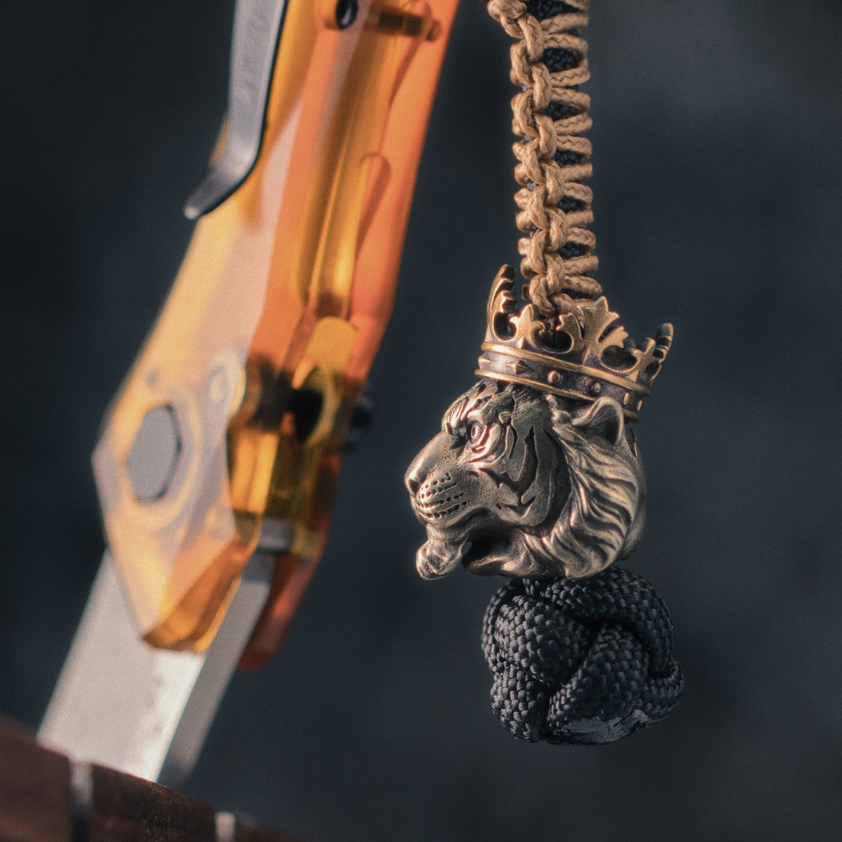 Tiger King paracord bead attached to a braided knife lanyard beside an EDC folding knife, with cupronickel and brass construction.