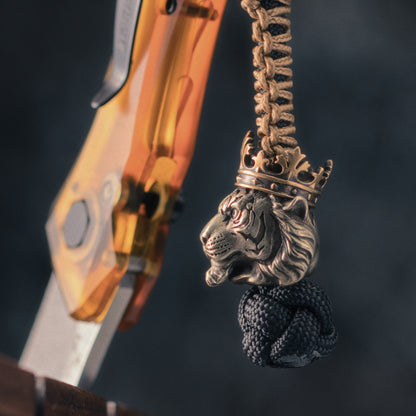Tiger King paracord bead attached to a braided knife lanyard beside an EDC folding knife, with cupronickel and brass construction.