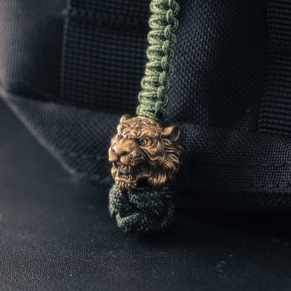 Three-quarter view of a brass tiger head paracord bead showing exposed fangs, carved facial stripes and hand-aged metal finish.